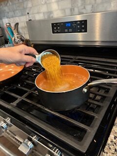 Pouring shredded cheddar cheese into a pot of tomato soup on the stove.