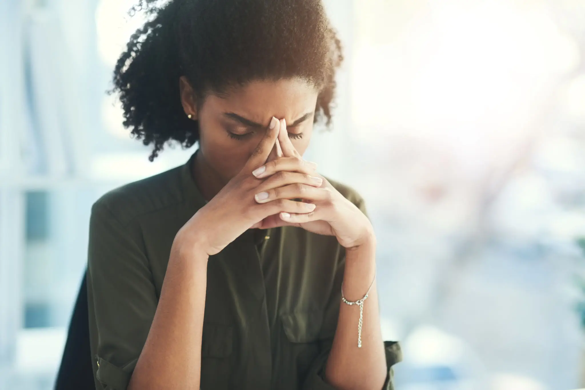 A woman looks stressed and thoughtful with hands clasped near her face.