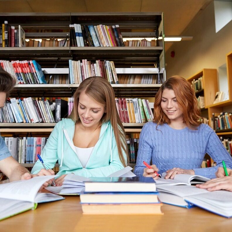 Three young women studying together in a library with books and notes.