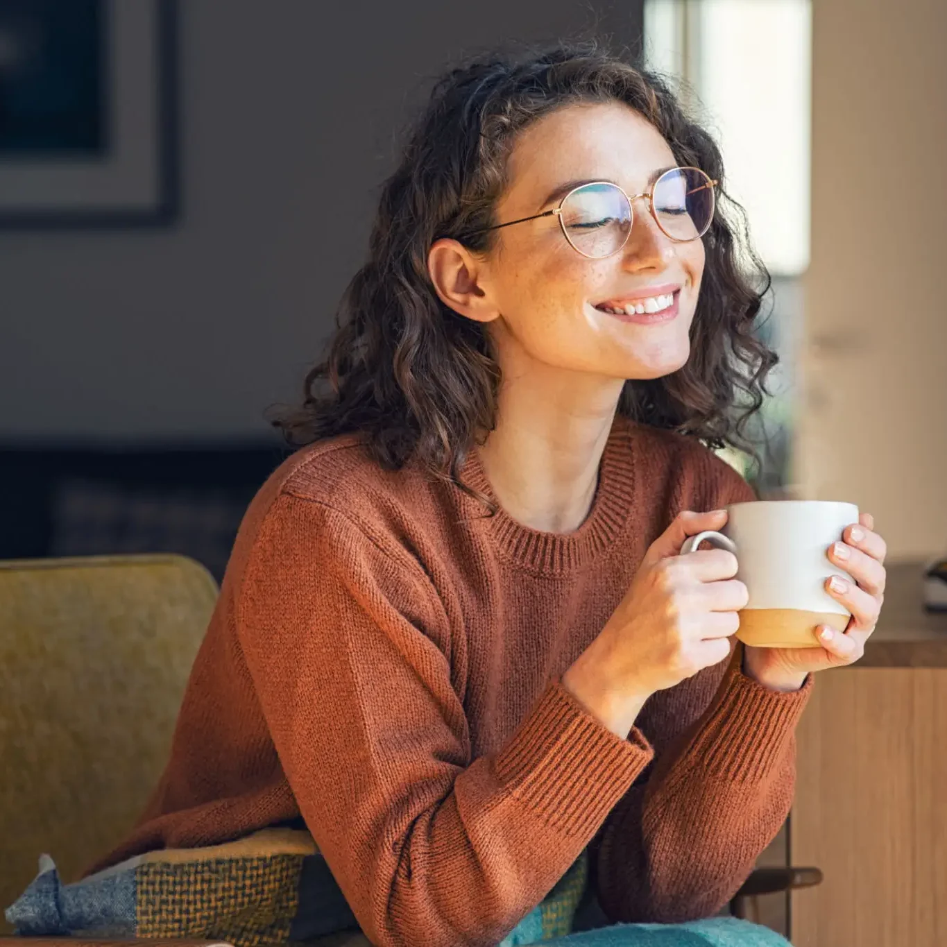 A woman in glasses smiling while holding a coffee mug indoors.